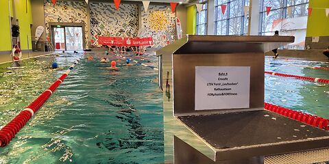 Am Startblock der Bahn 4 ist ein Blatt mit den Namen der Teams angebracht, die dort schwimmen: Crossfit, LTSV Forst "Laufsocken", Rathausteam, FORphysio&FORfittness. Auf der Bahn schwimmen mehrere Personen in beide Richtungen. Hinter dem Becken wird ein Banner der Sparkasse hochgehalten.