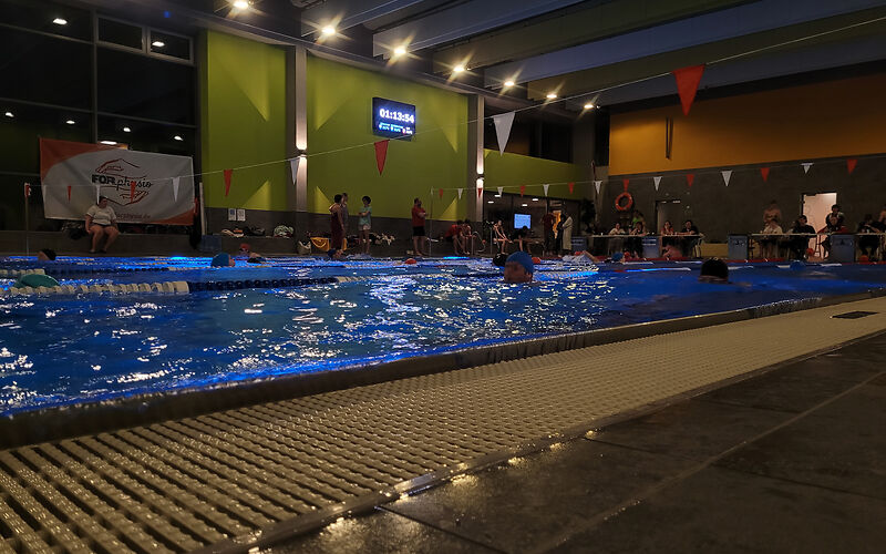 In der nur leicht beleuchteten Schwimmhalle schwimmen auf jeder Bahn mehrere Personen. Die Uhr an der Wand zeigt 01:13:54. Hinter dem Schwimmbecken stehen bzw. sitzen mehrere Teilnehemer, die pausieren. Vor dem Becken sitzen an Tischen pro Bahn jeweils 2 Leute.