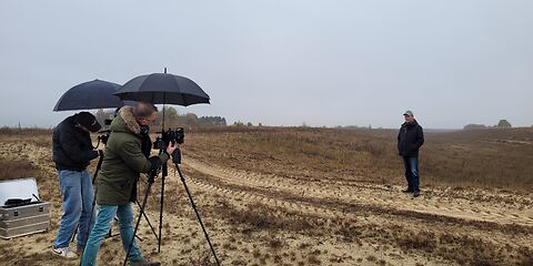 Auf einem Feld wird gearde das Interview gereht. Es regnet ein wenig und es ist sehr bewölkt. " Techniker stehen hinter 2 Kameras, die mit schwarzen Schirmen abdeckt sind.