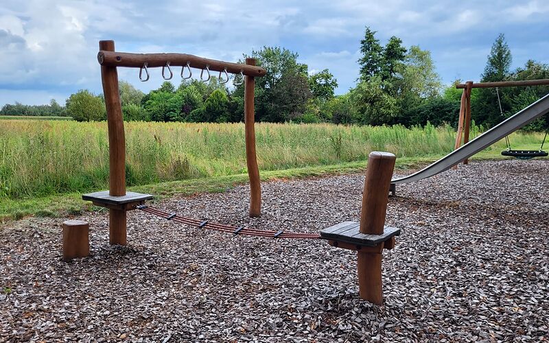 Spielgerät aus Holz mit drei verbundenen Seilen, die zwischen zwei Plattformen gespannt sind sowie einem waagerecht auf zwei Säulen liegenden Holzbalken, an dem mehrere Griffe befestigt sind.