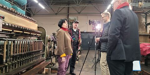 Im Museumsdepot filmen 2 Peronen ein Interview mit einer Dame. Diese steht vor einer Maschiene mit verschiedenen Spulen. Des Weiteren steht ein Mitarbeiter vom Museum mitdabei und hält 3 Spulen in den Farben galb, lila und blau in der Hand.