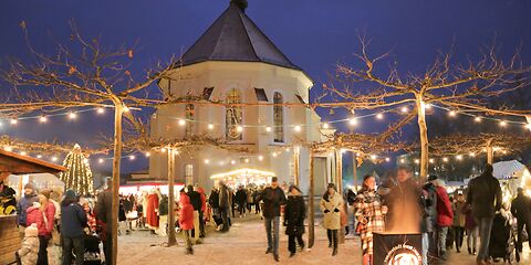 auf dem Forster Weihnachtsmarkt, mit Schneegestöber sind im Vordergrund unter dem mit Licherketten beleuchteten Platanenhain Menschen zu sehen, die an einer unter anderem an einer Feuerschale stehen. Im Hintergrund ist das Kirchenschiff der St. Nikolai zu sehen.