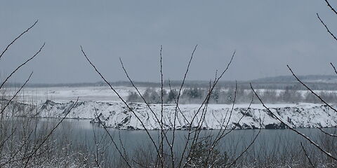 Nahaufnahme: Hinter verschneiten kahlen Ästen liegt der zugefrorene Klinger See. Im Hintergrund ist ein zugeschneiter Abhang zur Seeoberfläche und ein kleiner Wald in einer sonst leerstehenden Schneelandschaft zu sehen.