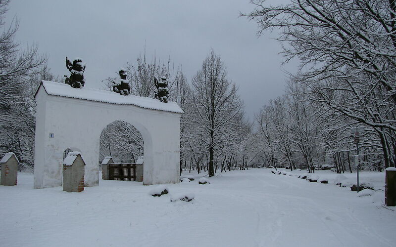 Ein weißer Torbogen, auf dem 3 Engel sitzen steht links im Bild. Mittig führ ein Weg in einen Wald. Die Landschaft ist komplett Schneebedeckt und am Himmel sind hellgraue Wolken.