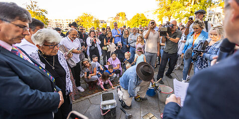 Künstler Gunter Demnig kniet auf dem Gehweg vor den eingesetzen Stolpersteinen, neben ihm stehen Eimer mit Beton. Um ihn herum stehen die Teilnehmer an der Veranstaltung, viele von ihnen machen Fotos. 