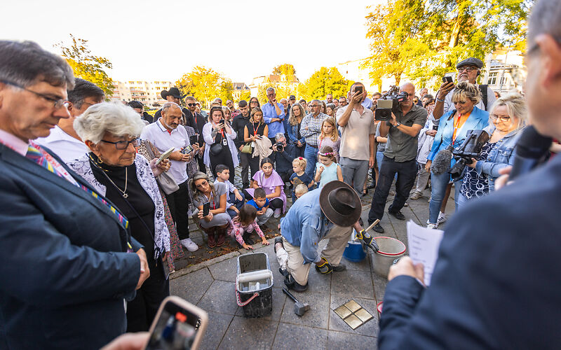 Künstler Gunter Demnig kniet auf dem Gehweg vor den eingesetzen Stolpersteinen, neben ihm stehen Eimer mit Beton. Um ihn herum stehen die Teilnehmer an der Veranstaltung, viele von ihnen machen Fotos. 