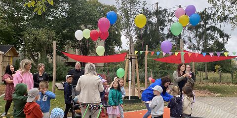 Kinder der Kita und Gäste auf der Einweihungsfeier. Es gibt Luftballons und eine Girlande als Deko. 