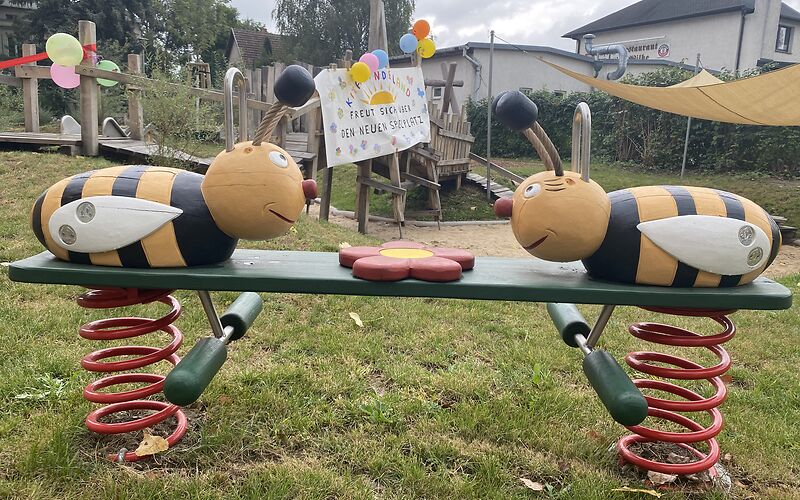 Eine Federwippe auf der an jeder Seite eine Biene aus Holz, die auf einem grünen Brett befestigt sind. In der Mitte ist eine Blume angebracht. Im Hintergrund befinden sich Luftballons und ein Plakat.