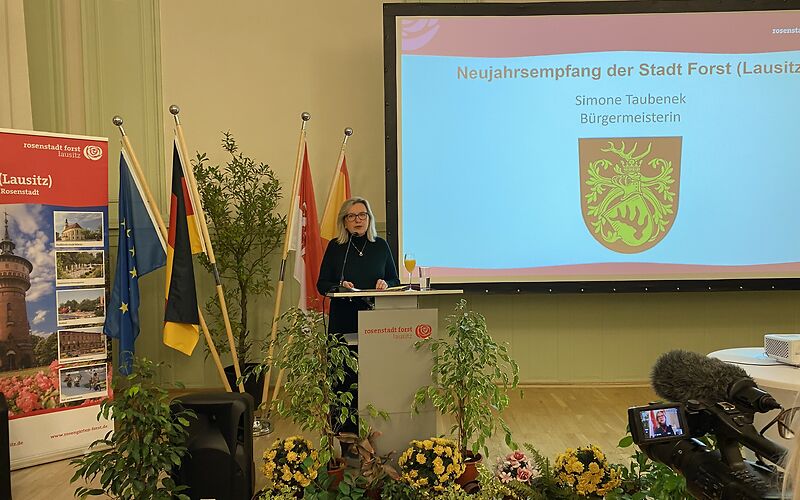 Bürgermeisterin Simone Taubenek spricht auf dem Neujahrsempfang und wird dabei gefilmt. Mittig auf der Bühne hängt eine große Tafeln, auf der eine Präsentation abgespielt wird. Links steht ein Banner der Stadt Forst (Lausitz).