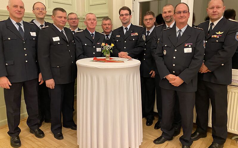 11 uniformierte Feuerwehrmänner stehen hinter einem weißen Tisch.