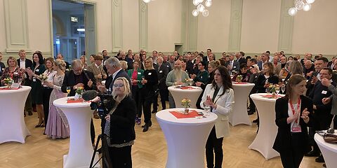 Blick in den Saal auf die Gäste des Neujahrsempfangs, welche an weißen Tischen stehen. Vorn steht eine Frau mit einer Kamera.