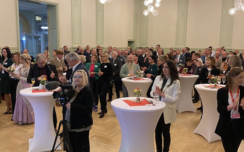 Blick in den Saal auf die Gäste des Neujahrsempfangs, welche an weißen Tischen stehen. Vorn steht eine Frau mit einer Kamera.