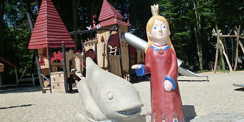 Eine Holzfigur einer prizessin mit blonden Haaren, blauen Augen und einem roten Kleid steht neben einer Statue eines Fisches. Im Hintergrund ist ein Klettergerüst, welches wie ein Schloss aussieht auf einem Sandplatz. 