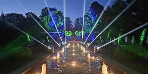 Die Wasserspiele im Rosengarten werden mit Lichter im Wasser und durch Lichstrahlen von Lasern mit weißen Strahlen und grünen Spiralen an beiden Seiten beleuchtet. Es ist kurz vorm Nachteinbruch und ein klarer, wolkenloser Himmel ist im Hintergrund. Die Wasserfontänen sprudeln leicht. 