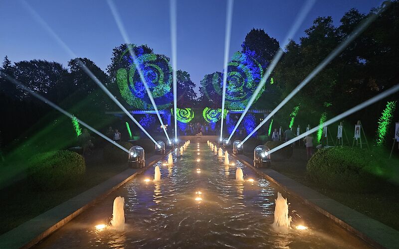 Die Wasserspiele im Rosengarten werden mit Lichter im Wasser und durch Lichstrahlen von Lasern mit weißen Strahlen und grünen Spiralen an beiden Seiten beleuchtet. Es ist kurz vorm Nachteinbruch und ein klarer, wolkenloser Himmel ist im Hintergrund. Die Wasserfontänen sprudeln leicht. 