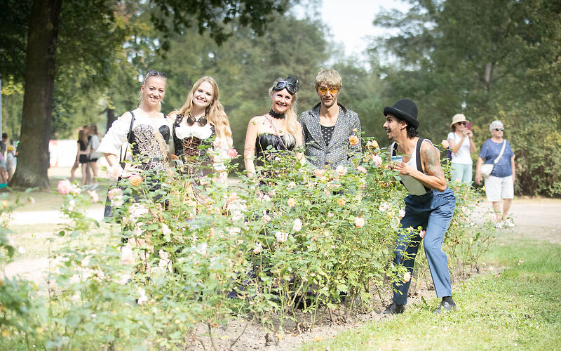 Mehrere Besucher des Festivals hinter einem Rosenbeet im Rosengarten, 5 davon zentral im Bild und weitere im Hintergrund. 