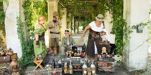 Im Steampunk-Stil gestaltete Koffer, Jetpacks und Maschinen, dahinter stehen bzw. sitzen mehrere Personen.