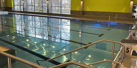 Das Schwimmerbecken mit Blick von der Treppe, die in das Becken führt, aus. Das Becken hat 5 Bahnen, die auf dem Boden durch dicke schwarze Linien getrennt werden.