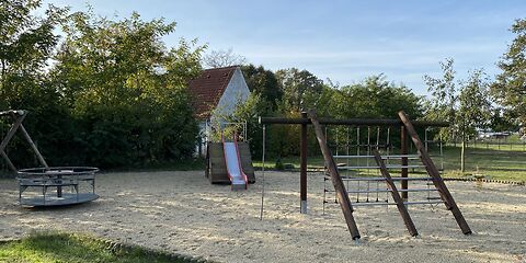 Ein Holzklettergerüst steht im vorderen rechten Bereich. Dahinter befindet sich eine Rutsche mit roten Seiten auf einem Holzgestell. Links im Bild ist ein rundes Karussel. 