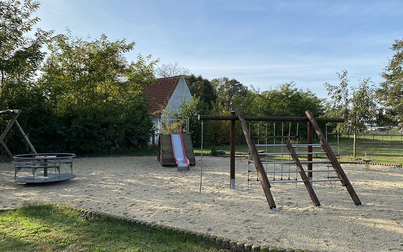 Ein Holzklettergerüst steht im vorderen rechten Bereich. Dahinter befindet sich eine Rutsche mit roten Seiten auf einem Holzgestell. Links im Bild ist ein rundes Karussel. 