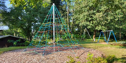 Ein Kletterturm mit bunten Seilen und einer Metallstange in der Mitte, im Hintergrund ist eine Doppelschaukel.