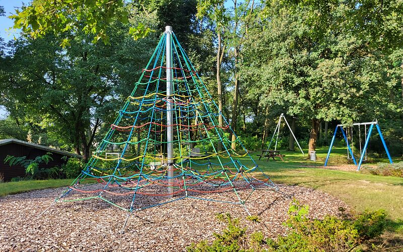 Ein Kletterturm mit bunten Seilen und einer Metallstange in der Mitte, im Hintergrund ist eine Doppelschaukel.