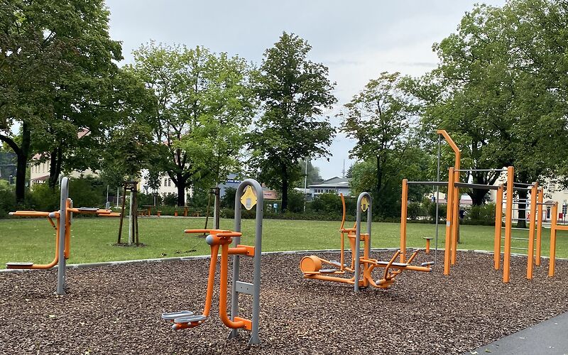 Sportgeräte auf dem Spielplatz, gestaltet in den farben orange und grau sin im Vodergrund, darunter Arm-Bauchmuskeltrainer und Crisstrainer.