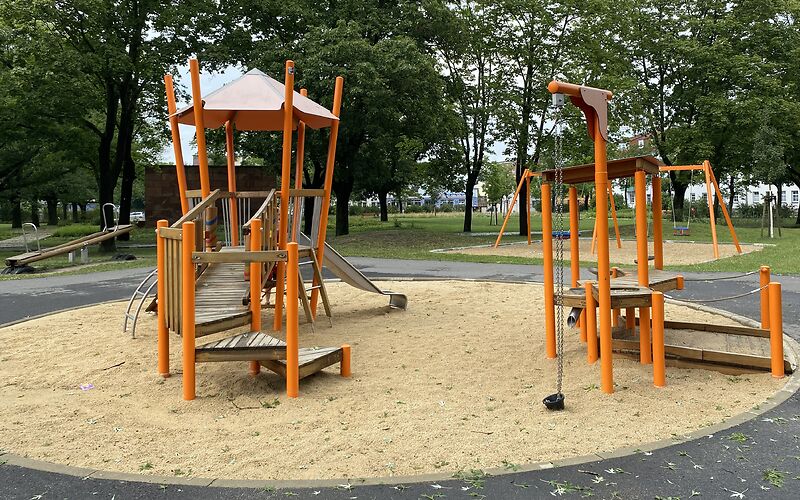 Auf einem Sandplatz steht in Turm mit Geüst mit Dach aus Orangenen Stangen und einem Aufstieg aus Holz. An Turm ist eine silberne Rutsche.