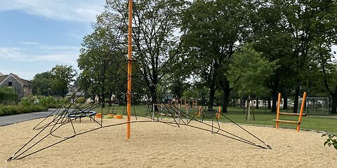 Auf einem Sandplatz steht ein orangener Mast, um welchem ein Netzt aus schwarzen Seilen gespannt ist, welches unten breit ist und nach oben hin schmaler wird. 