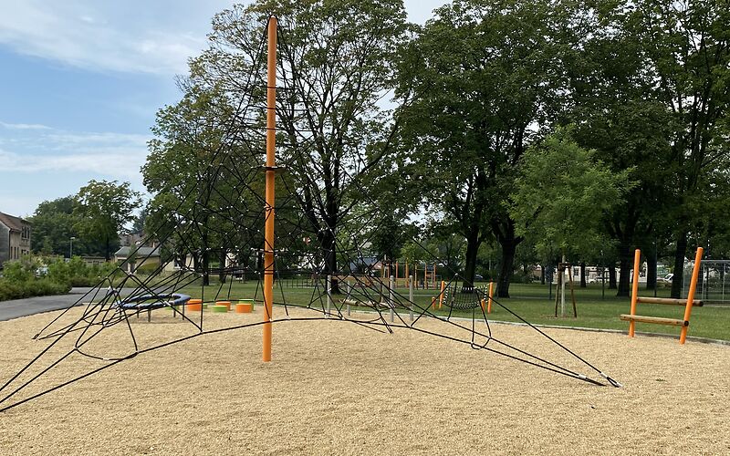 Auf einem Sandplatz steht ein orangener Mast, um welchem ein Netzt aus schwarzen Seilen gespannt ist, welches unten breit ist und nach oben hin schmaler wird. 