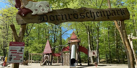 Zwischen zwei im Boden befestigen Holzpfälen ist ein Schild mit der Aufschrift "Dornröschenpark" angebracht. Dies ist der Eingang zum Spielplatz. Im Hintergrund befindet sich das Klettergrüst mit einer Rutsche.