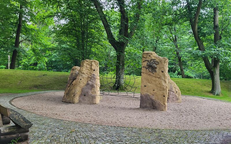 Ein Kletternetz aus Seilen, welches zwischen zwei Felsen gespannt ist. Um das Gerüst herum ist eine runde Fläche aus Sand, dahinter befinden sich Wiese und Bäume. Im Vorderung ist gepflastert.