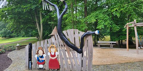 Ein hölzernen Zaun auf dem eine Prizessin und ein König befestigt sind. Statt den Gesichtern sich Löcher eingebaut, wo Kinder ihren Kopf durchstecken können. Im Hintergrund sind Bäume auf einer WIese.