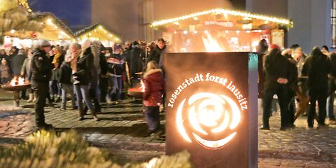 Vorn leuchtet das Rosen-Logo der Stadt Forst mit dem Schriftzug "rosenstadt forst lausitz", im Hintergrund sind zahlreiche Besucher auf dem Weihnachtsmarkt an Tischen und Feuerschalen sowie mehrere Hütten auf dem Weihnachtsmarkt zu sehen.