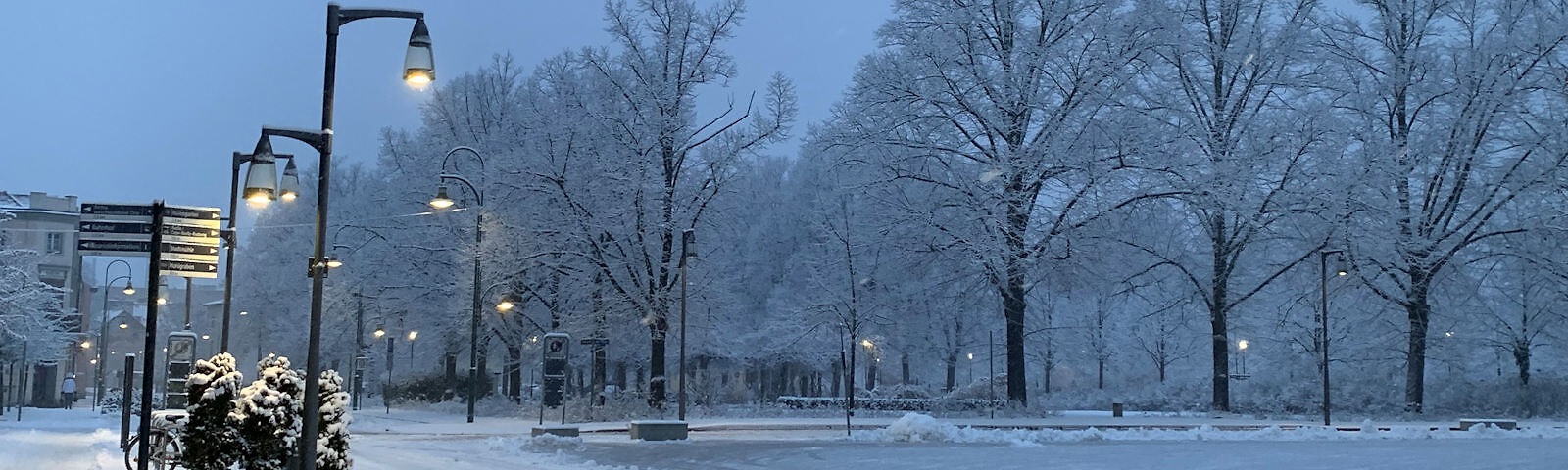 Blick auf den verschneiten Lindenplatz, es liegt eine weiße Schneedecke auf dem Boden, die Äste der Bäume sind mit Schnee bedeckt, die Straßenlaternen leuchten bereits