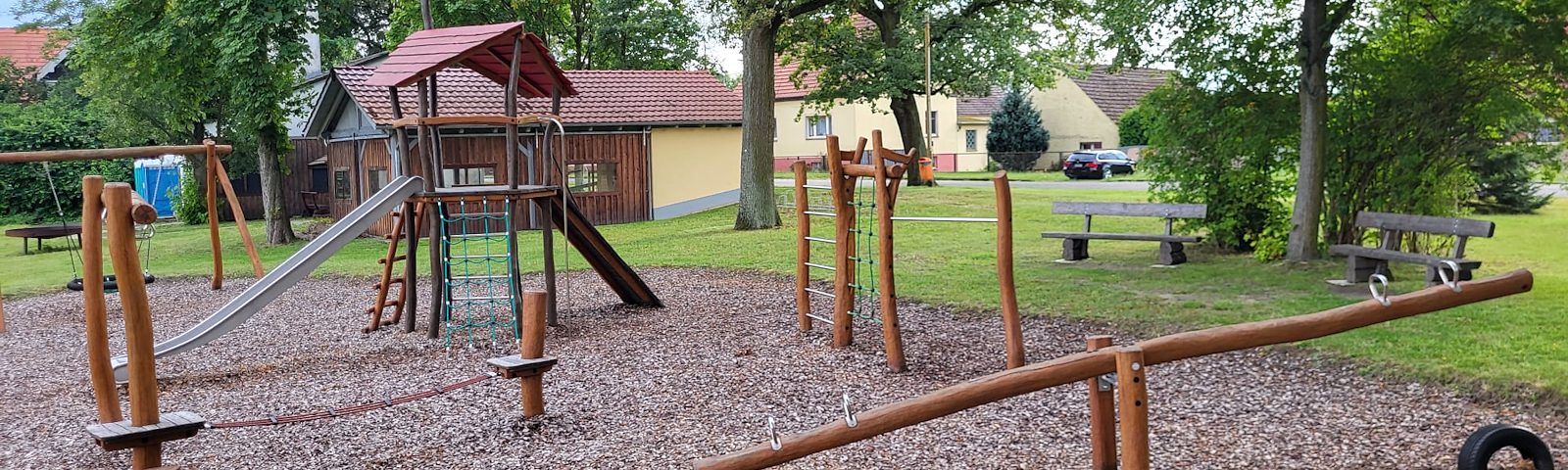 Ein Kletterturm mit Rutsche, mehrere Kletterstangen und eine Wippe, im Hintergrund steht eine Schaukel, zwei Bänke rechts neben dem Spielplatz