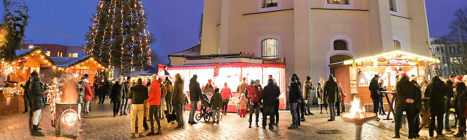 Vor der Stadtkirche stehen mehrere Stände des Forster Weihnachtsmarktes, davor stehen die Besucher an Stehtischen und zwei Feuerschalen versammelt. Im Hintergrund ist der beleuchtete Weihanchtsbaum neben der Stadtkirche zu sehen.