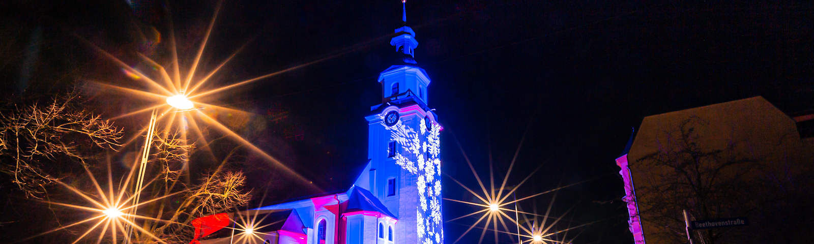 Die Stadtkirche Forst wird durch Lichter angestrahlt, sodass auf der Turmseite Schneeflocken zu sehen sind. Der Himmel dahinter ist dunkel, vorn leuchten 5 Straßenlatern.