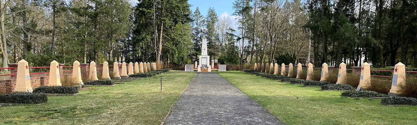 Ein geflasterter Weg, an dessen Seiten mehrere Grabsteine in einer Reihe auf einem Wiesenstreifen stehen. Der Weg führt zu einem großen Denkmal. Ringsherum stehen Bäume.zu sehen.