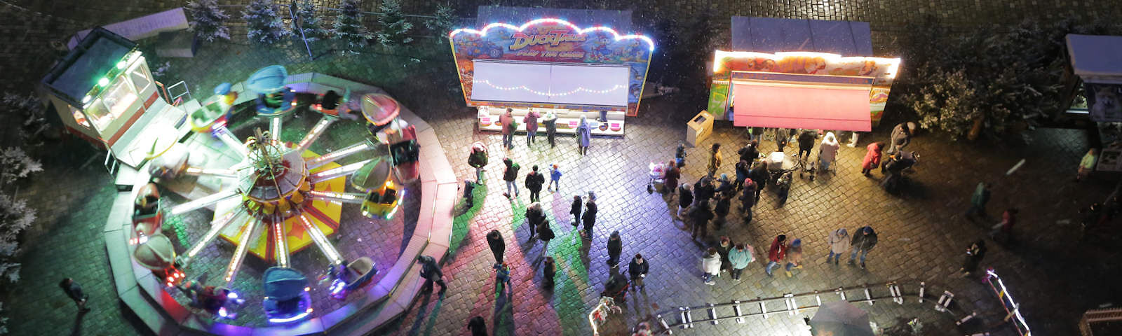 Blick von oben auf den Forster Weihnachtsmarkt: links ist ein Fahrgeschäft im Betrieb, vor mehreren Ständen stehen Personen, die Schienen eine Kindereisenbahn sind zu sehen. Der Platz und die Umgebung sind beleuchtet,