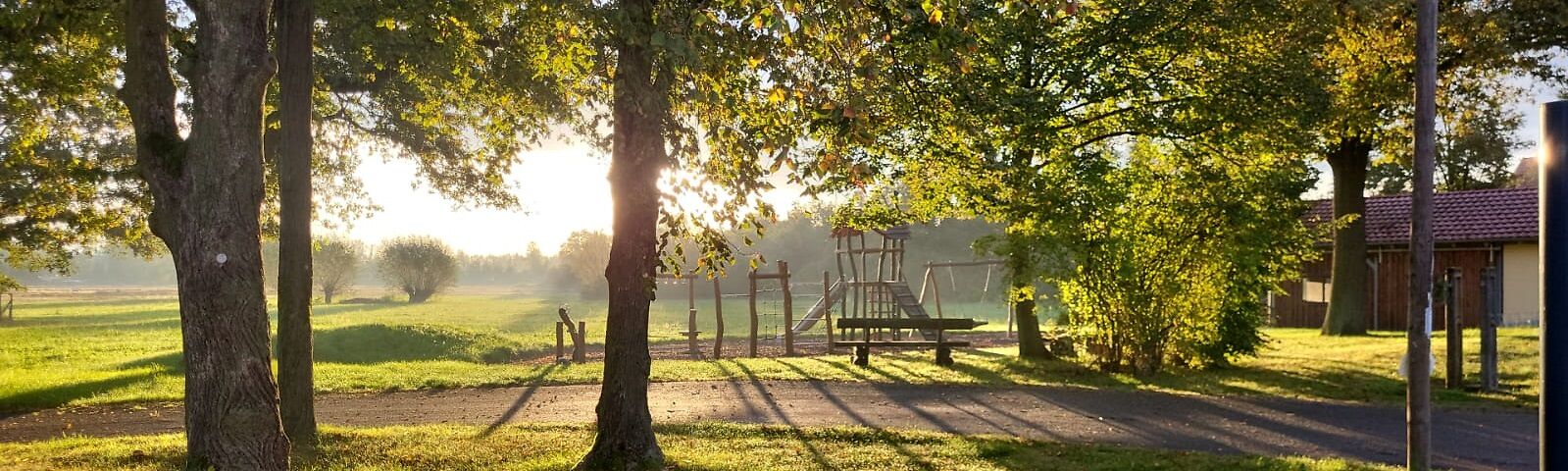 Zwischen Bäumen auf einer Wiese ist im Sonnenaufgang ein Spielplatz zu sehen. Vor dem Spielplatz verläuft ein Weg. Rechts steht ein Haus.